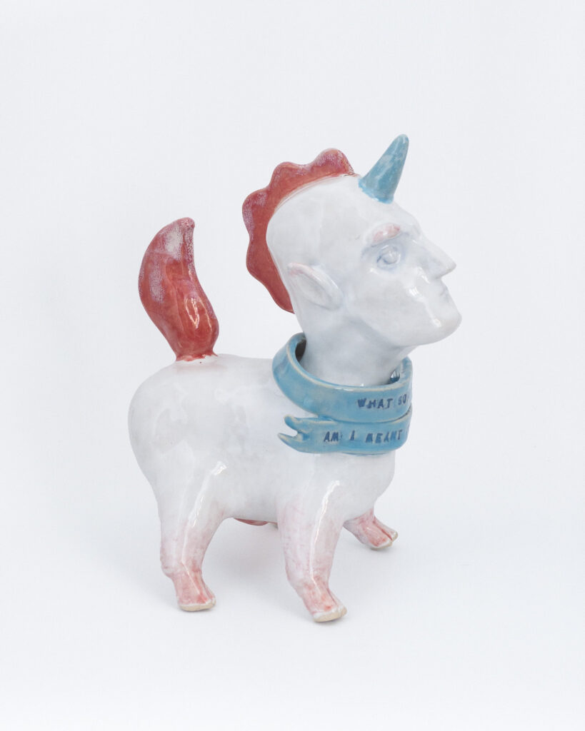 A ceramic unicorn creature with a squat body and a human head, wearing a scarf that reads "What sort of creature am I meant to be?", side view