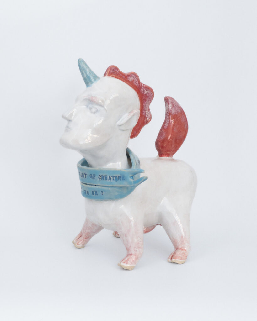 A ceramic unicorn creature with a squat body and a human head, wearing a scarf that reads "What sort of creature am I meant to be?"