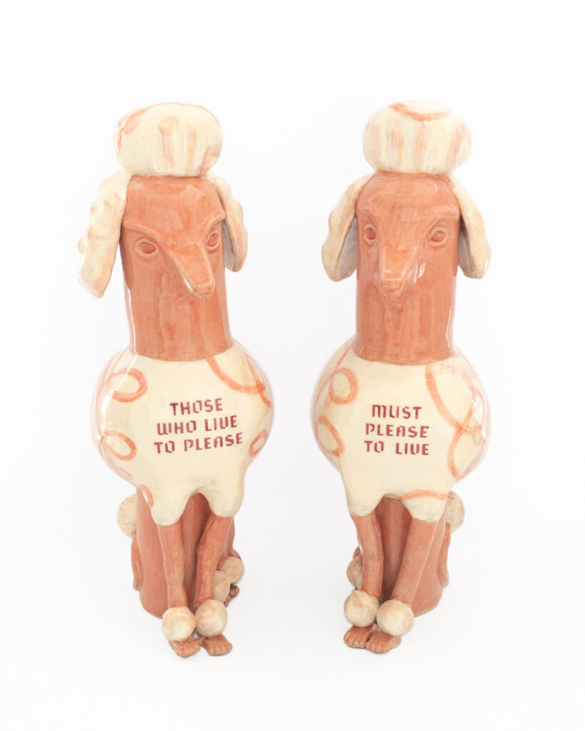 Pair of ceramic poodles in pink glazes and swirly designs with the text "Those Who Live to Please" and "Must Please to Live" on their chests