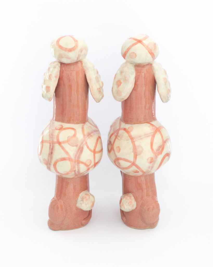 Pair of ceramic poodles in pink glazes and swirly designs with the text "Those Who Live to Please" and "Must Please to Live" on their chests, from behind