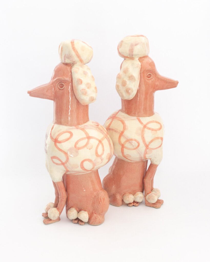Pair of ceramic poodles in pink glazes and swirly designs with the text "Those Who Live to Please" and "Must Please to Live" on their chests, turned to show their sides
