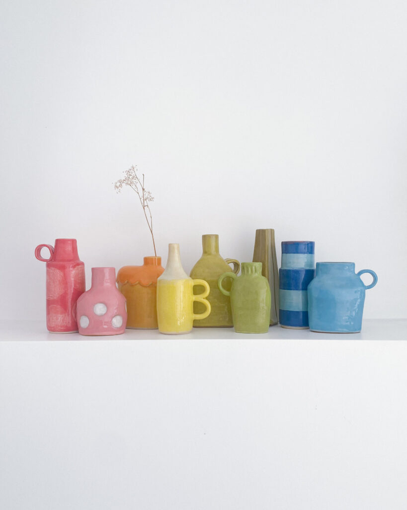 A row of small ceramic bud vases, all different shapes and styles, in the colours of the rainbow