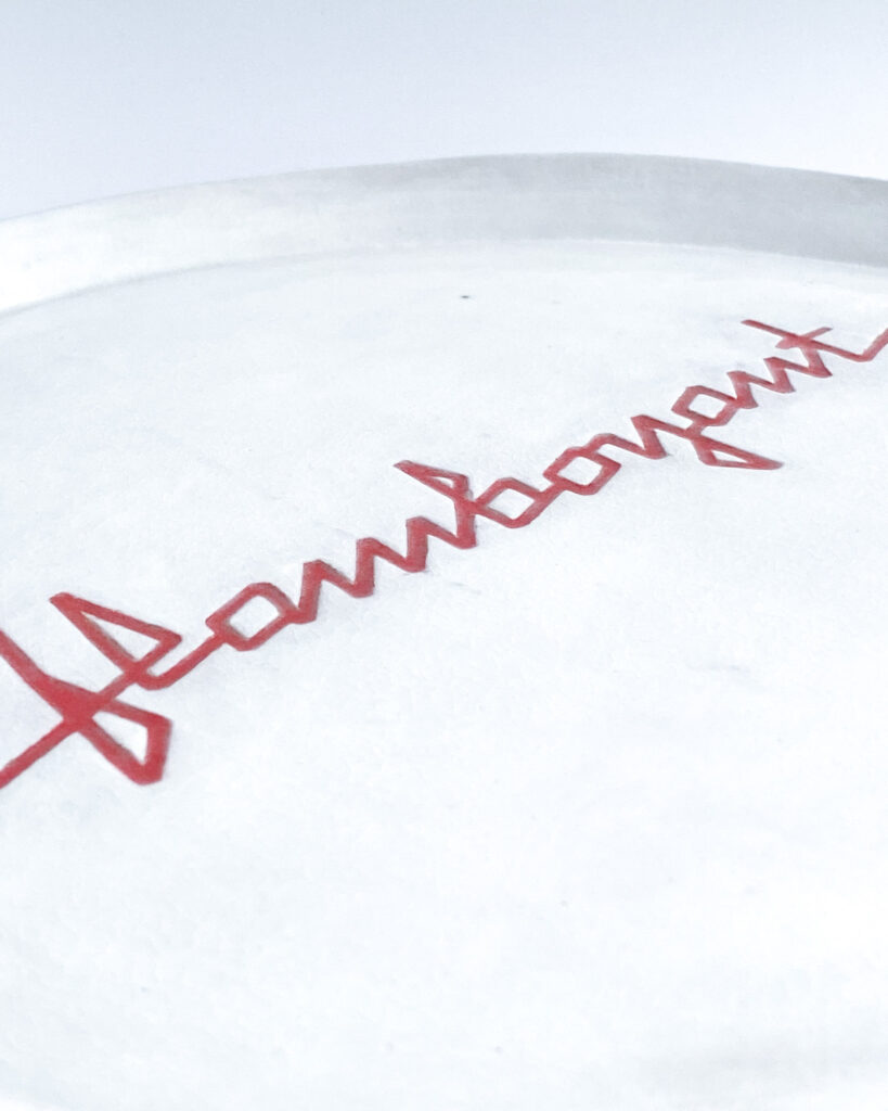 White ceramic plate with "flamboyant" carved across the surface in angular red ribbon letters, close up