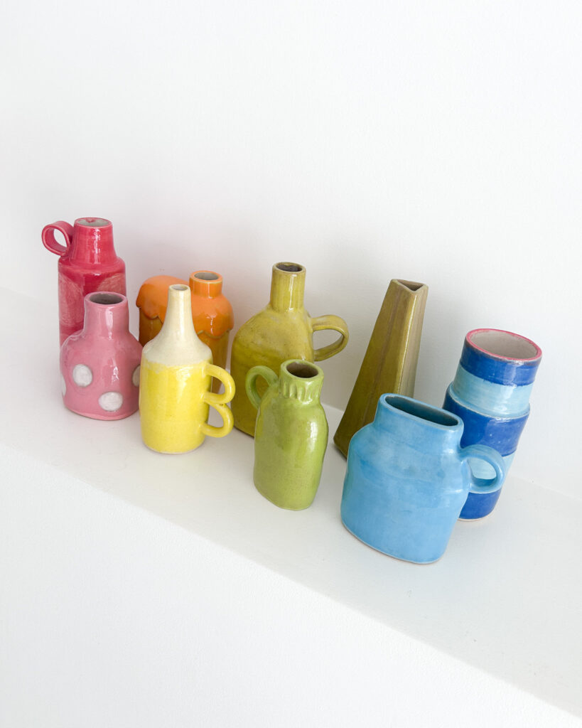 A row of small ceramic bud vases, all different shapes and styles, in the colours of the rainbow