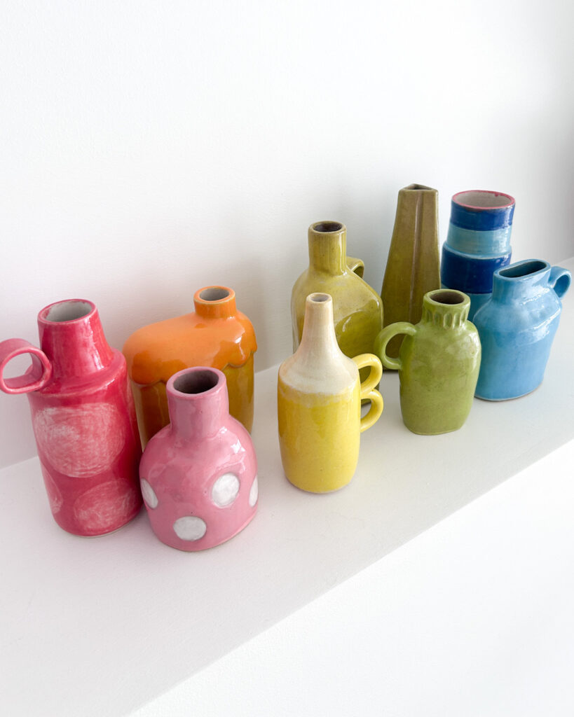A row of small ceramic bud vases, all different shapes and styles, in the colours of the rainbow