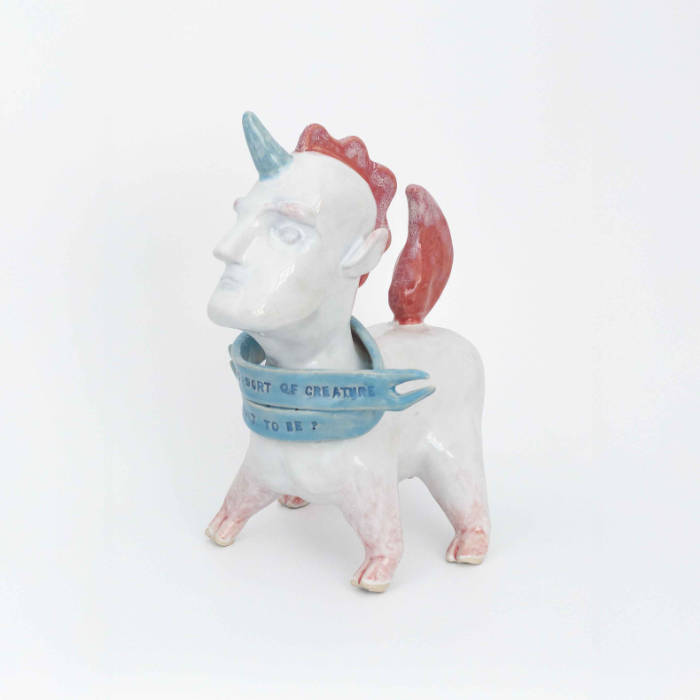 A ceramic unicorn creature with a squat body and a human head, wearing a scarf that reads "What sort of creature am I meant to be?"